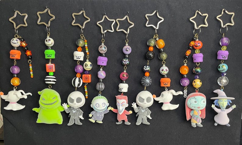 May include: A collection of Halloween-themed keychains featuring characters from The Nightmare Before Christmas. Each keychain has a star-shaped top and a beaded chain with character charms, including Jack Skellington, Sally, and Zero.