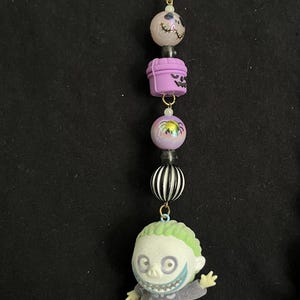 May include: A keychain featuring a star-shaped charm and a series of decorative beads. The beads include a skull, a purple container, a round bead with a spider, a black and white striped bead, and a figure of a character with green hair.
