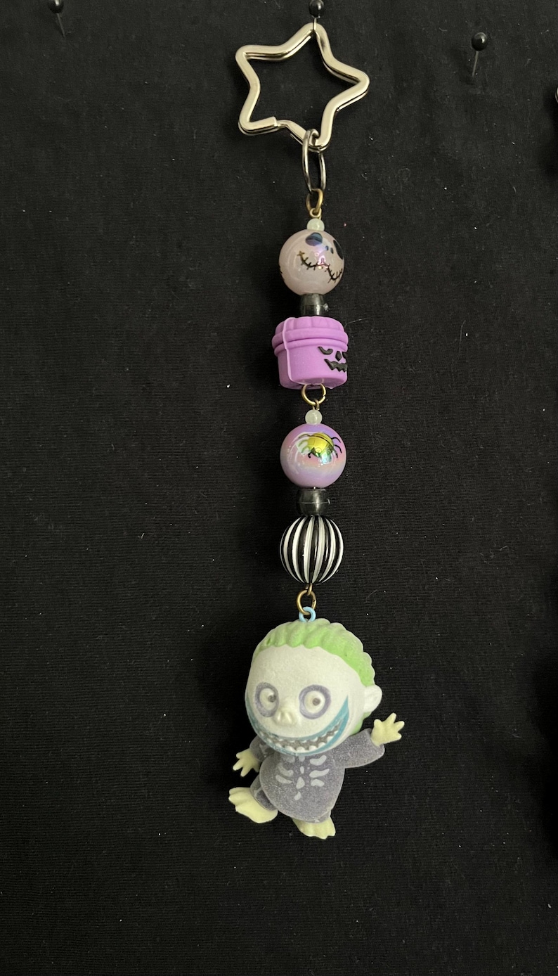May include: A keychain featuring a star-shaped charm and a series of decorative beads. The beads include a skull, a purple container, a round bead with a spider, a black and white striped bead, and a figure of a character with green hair.