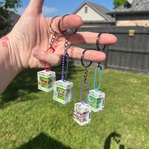 May include: Four keychains with small, clear boxes containing cartoon character figures. Each box is attached to a black metal ring with a chain of beads in red, purple, and blue. The keychains are held up against a background of green grass and a blue sky.