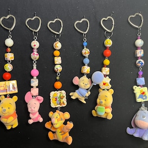 May include: A selection of keyrings featuring Winnie the Pooh characters. Each keyring has a heart-shaped loop, colourful beads, and a character charm. Characters include Winnie the Pooh, Piglet, Tigger, Eeyore, and Roo.