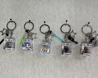 May include: Five Star Wars themed keychains featuring miniature Funko Pop figures in clear display boxes. Each keychain includes a metal ring, a star-shaped charm, and a specific accessory like a lightsaber, blaster, or dumbbell charm.