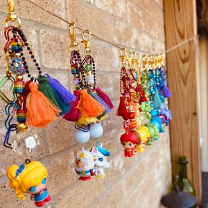 May include: A collection of colorful keychains hanging on a string. Each keychain features a gold clasp, beads, tassels, and a small figurine. The figurines include a superhero, a sheep, a unicorn, and other characters. The keychains are displayed against a brick wall.