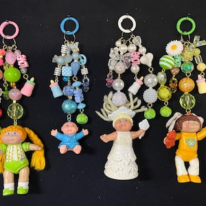 Könnte beinhalten: Vier Schlüsselanhänger mit Cabbage Patch Kids-Puppen. Jeder Schlüsselanhänger hat ein anderes Farbschema aus Perlen, Charms und einer Puppe. Die Puppen tragen verschiedene Outfits und haben unterschiedliche Haarfarben. Die Schlüsselanhänger sind an einem Metallring befestigt.