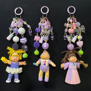 May include: Three Cabbage Patch Kids keychains with purple, white, and gold accents. Each keychain features a doll with a unique outfit and a cascade of beads, bows, and charms in shades of purple and pink. The keychains have a ring at the top.