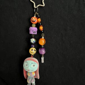 May include: A Halloween-themed keychain featuring a star-shaped clasp. The keychain includes orange and purple beads, a Sally figure, and pumpkin and coffin charms. The word "Boo" is written on a charm.