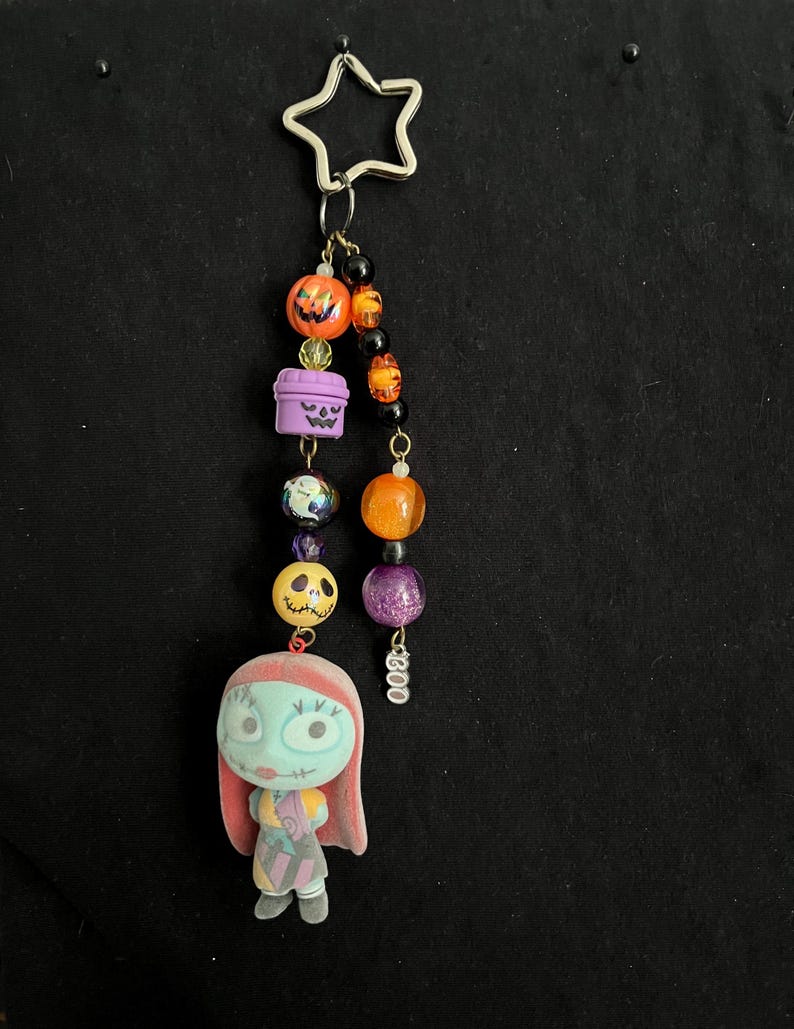 May include: A Halloween-themed keychain featuring a star-shaped clasp. The keychain includes orange and purple beads, a Sally figure, and pumpkin and coffin charms. The word "Boo" is written on a charm.