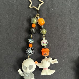 May include: A Halloween-themed keychain featuring Jack Skellington and Zero from The Nightmare Before Christmas. The keychain includes a star-shaped clasp, beads in orange, black, and green, and a spiderweb charm. The figures are white and gray.