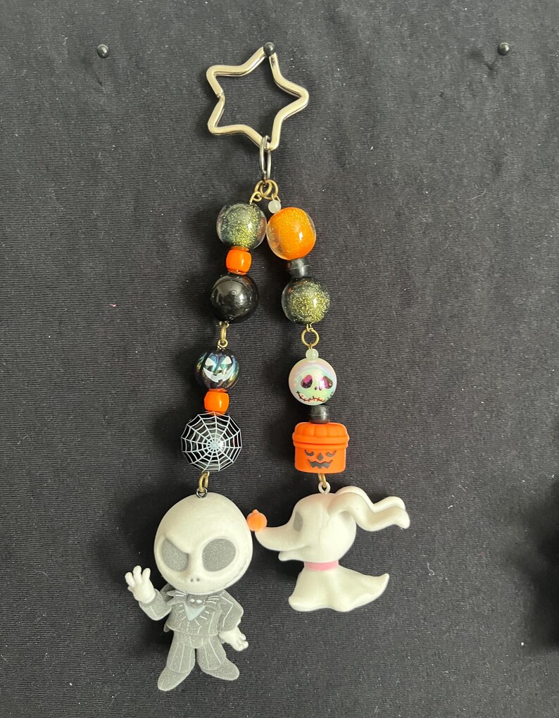 May include: A Halloween-themed keychain featuring Jack Skellington and Zero from The Nightmare Before Christmas. The keychain includes a star-shaped clasp, beads in orange, black, and green, and a spiderweb charm. The figures are white and gray.