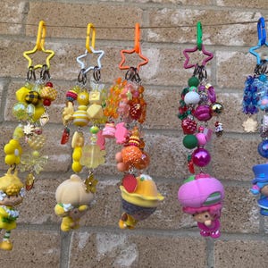 Puede incluir: Cinco llaveros coloridos con clips en forma de estrella. Cada llavero presenta un diseño único con cuentas, abalorios y una pequeña figura. Los llaveros son en tonos de amarillo, naranja, rosa y azul, con varias formas y texturas.