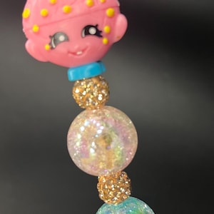 May include: A pink and yellow character charm with a blue headband, attached to a beaded bracelet with iridescent beads and gold accents.