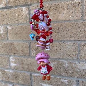 May include: A red plastic clip holds a charm with red, pink, and white beads, and miniature charms. The charms include a Strawberry Shortcake figurine, a cake, and other food-themed trinkets. The background is a brick wall.