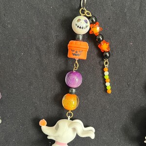 May include: A Halloween-themed charm featuring a star-shaped top, a smiling skull bead, an orange jack-o'-lantern, purple and orange beads, and a white Zero dog figurine. The charm is attached to a metal clasp.