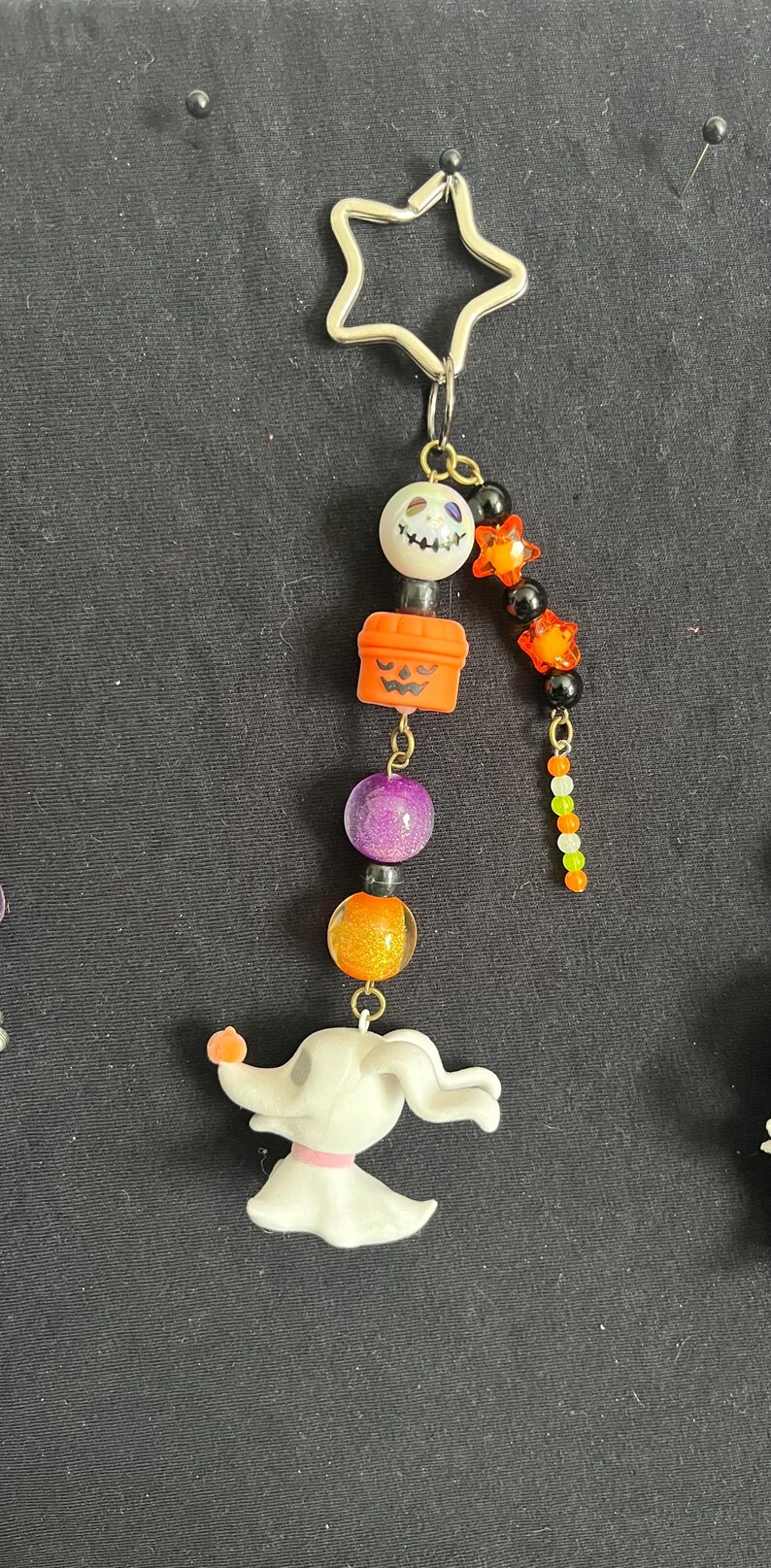 May include: A Halloween-themed charm featuring a star-shaped top, a smiling skull bead, an orange jack-o'-lantern, purple and orange beads, and a white Zero dog figurine. The charm is attached to a metal clasp.