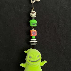 May include: A Halloween-themed keychain featuring a star-shaped top, a series of decorative beads, and a green Oogie Boogie figure. The beads include a skull, a jack-o'-lantern, a spiderweb, and a striped ball. The keychain is suspended against a black background.