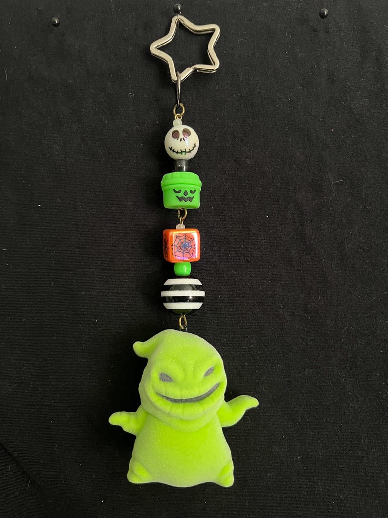 May include: A Halloween-themed keychain featuring a star-shaped top, a series of decorative beads, and a green Oogie Boogie figure. The beads include a skull, a jack-o'-lantern, a spiderweb, and a striped ball. The keychain is suspended against a black background.