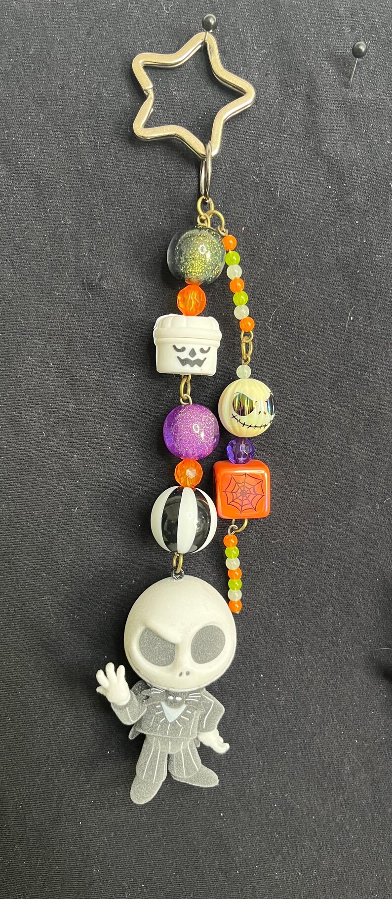 May include: A Halloween-themed keychain featuring a star-shaped top and a series of decorative beads. The beads include a green glitter ball, a white pumpkin, a purple glitter ball, a black and white ball, and a Jack Skellington figure.