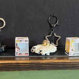 Op de afbeelding: Drie Funko Pop sleutelhangers met personages in voertuigen: een zwarte Batmobile met Batman, een witte Millennium Falcon met Han Solo en een perzikkleurige landspeeder met Luke Skywalker, elk vergezeld door het bijbehorende kleine doosje.