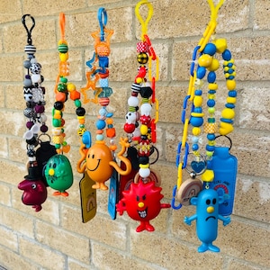 Puede incluir: Una colección de llaveros con cuentas de colores con varios dijes, incluyendo personajes de dibujos animados y formas. Los llaveros cuelgan contra una pared de ladrillos. Las cuentas son de varios colores.