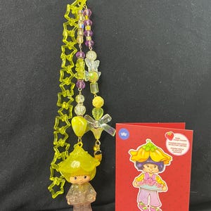 May include: A yellow and purple beaded keychain with a small Almond Tea figurine and a red card featuring a cartoon character holding a tray. The keychain has star-shaped and round beads.