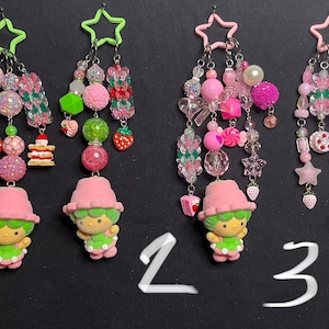 May include: Four pairs of dangle earrings with a variety of pink beads, charms, and a small figurine. The earrings feature star-shaped and green or pink star-shaped tops. The figurines are wearing pink hats and green dresses.