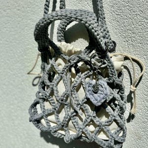 May include: A grey crocheted bag with a white lining and a heart-shaped charm. The bag has a drawstring closure and a long strap.