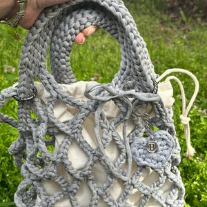 May include: A grey crocheted tote bag with a white lining and a grey crocheted heart charm with a silver letter "B" on it.