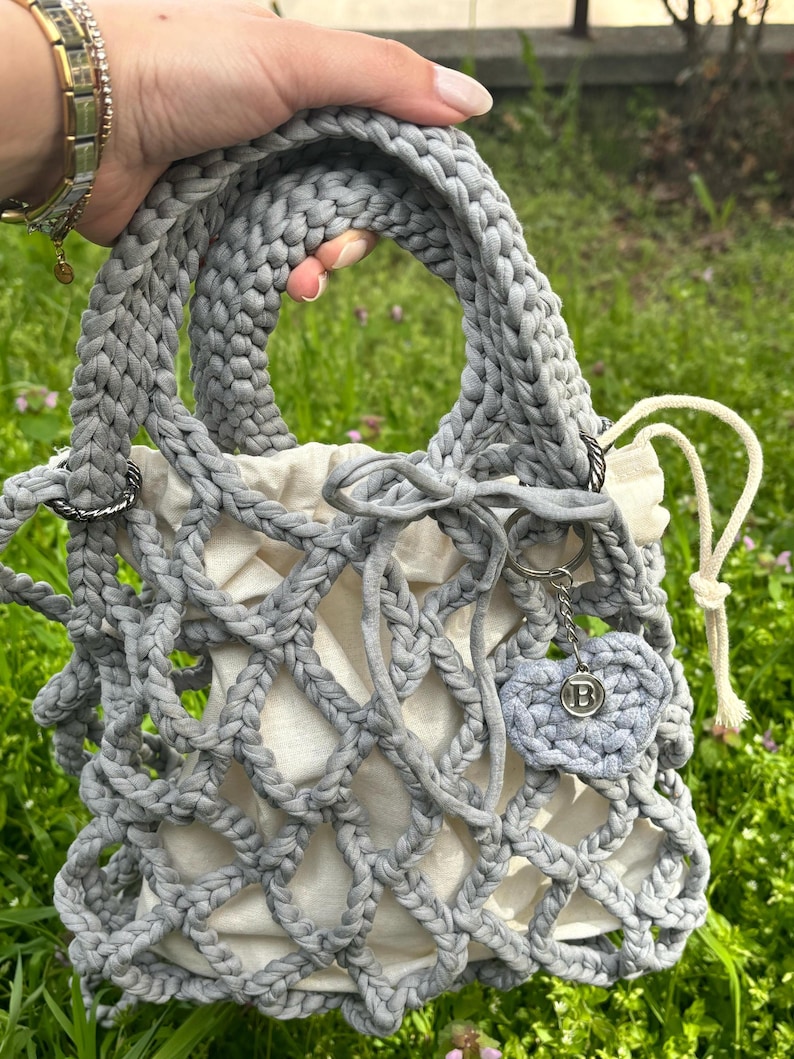 May include: A grey crocheted tote bag with a white lining and a grey crocheted heart charm with a silver letter "B" on it.