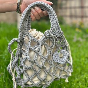 May include: A grey crocheted bag with a white fabric lining and a small grey crocheted heart charm with a metal 'B' charm. The bag has a drawstring closure and a long grey crocheted strap.