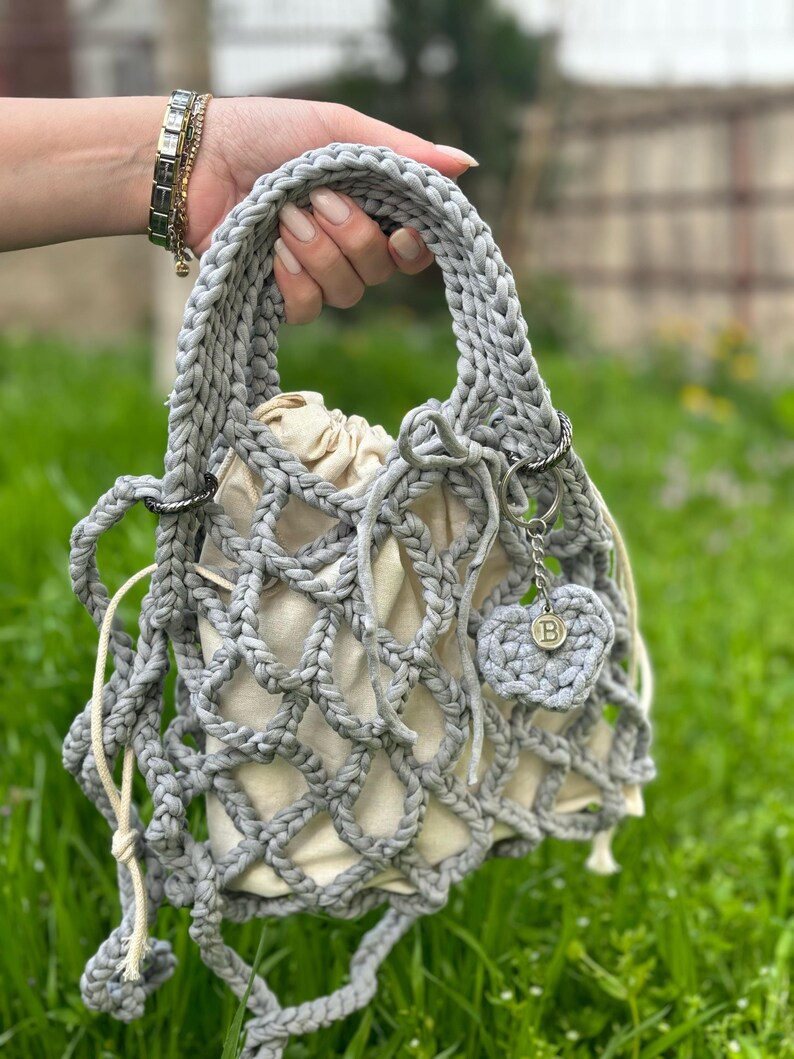 May include: A grey crocheted bag with a white fabric lining and a small grey crocheted heart charm with a metal 'B' charm. The bag has a drawstring closure and a long grey crocheted strap.