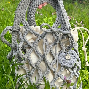 May include: A grey crocheted tote bag with a white mesh lining and a grey crocheted charm with the letter "B" hanging from the handle.