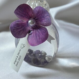 May include: A clear glass bottle filled with small stones, topped with a purple flower and a pearl. A tag with the text "Angel & David June 2025" hangs from the bottle. The bottle is likely a wedding favor or decorative item.