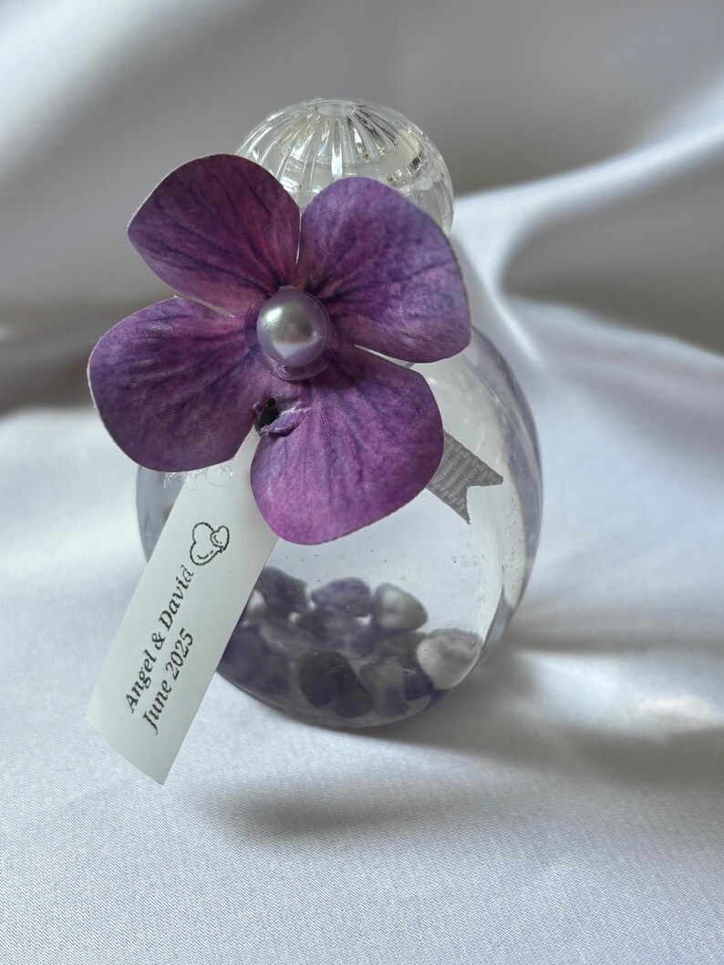 May include: A clear glass bottle filled with small stones, topped with a purple flower and a pearl. A tag with the text "Angel & David June 2025" hangs from the bottle. The bottle is likely a wedding favor or decorative item.