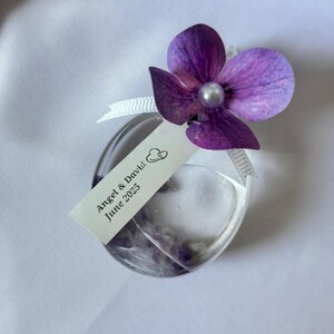 May include: A clear, heart-shaped glass bottle with a purple flower and a pearl bead. The bottle has a white tag that reads "Angel & David June 2025". The bottle is tied with a white ribbon.