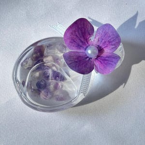 May include: A clear, oval-shaped container filled with small, purple and clear stones, topped with a vibrant purple flower with a pearl center. A light blue ribbon is attached to the container. The item is set against a white background.