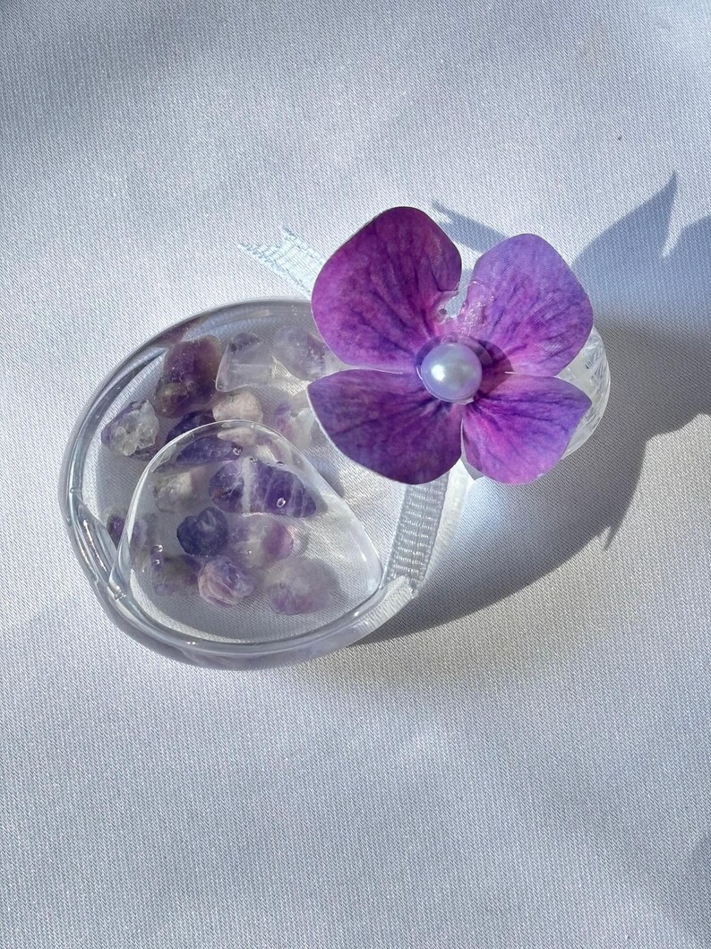May include: A clear, oval-shaped container filled with small, purple and clear stones, topped with a vibrant purple flower with a pearl center. A light blue ribbon is attached to the container. The item is set against a white background.