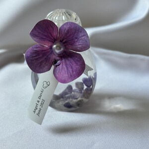 May include: A clear glass ornament filled with small purple stones, adorned with a large purple flower and a pearl center. A small tag reads "Angel & David June 2025". The ornament is set against a white background.