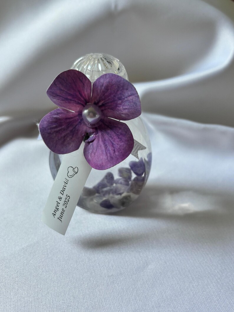 May include: A clear glass ornament filled with small purple stones, adorned with a large purple flower and a pearl center. A small tag reads "Angel & David June 2025". The ornament is set against a white background.
