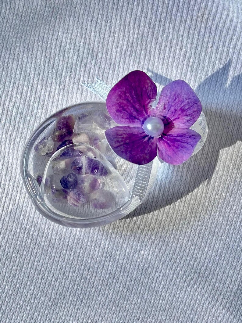 May include: A clear, oval-shaped container filled with small, purple and white stones. A vibrant purple flower with a pearl center is attached to the container with a light blue ribbon. The container sits on a white, textured surface.