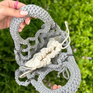 May include: A grey crocheted tote bag with black metal rings. The bag is open and has a white fabric lining.