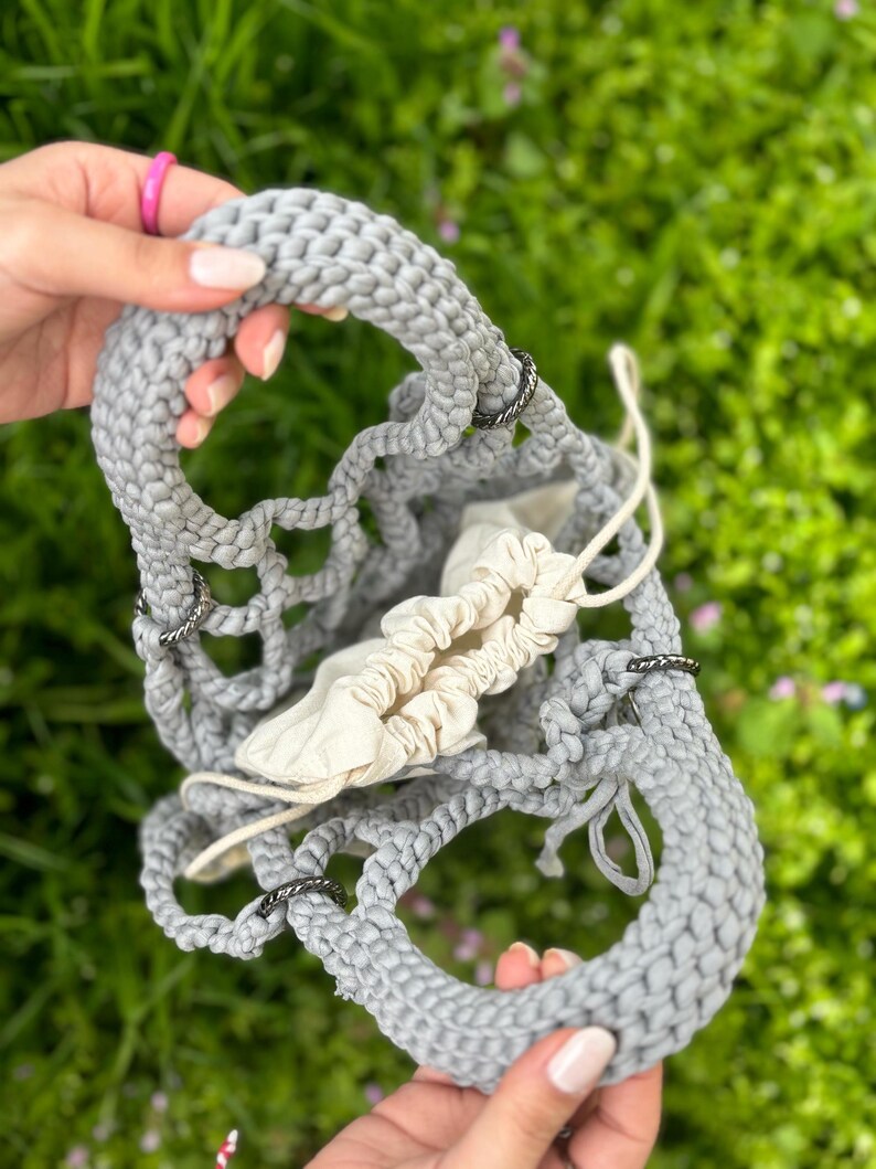 May include: A grey crocheted tote bag with black metal rings. The bag is open and has a white fabric lining.