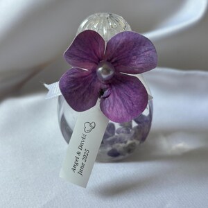 May include: A small, clear glass bottle filled with purple stones, topped with a clear, textured stopper. A purple flower with a pearl center is attached. A small tag reads "Angel & David June 2025".