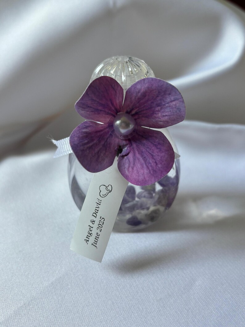 May include: A small, clear glass bottle filled with purple stones, topped with a clear, textured stopper. A purple flower with a pearl center is attached. A small tag reads "Angel & David June 2025".