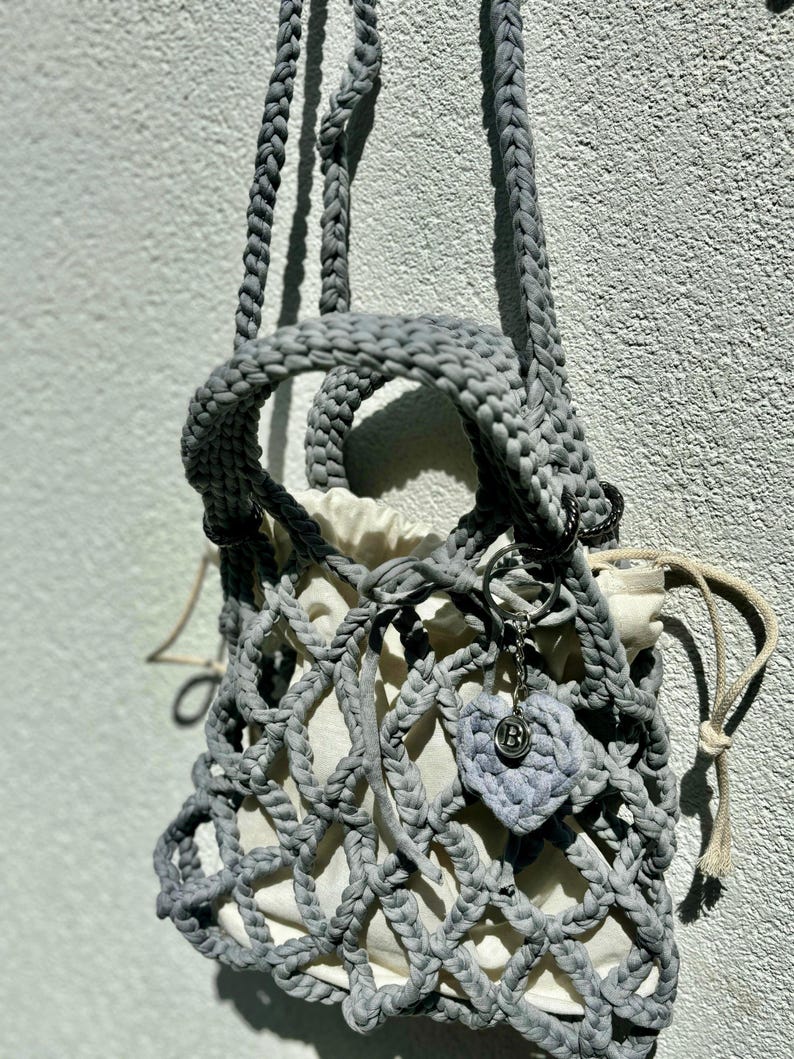May include: A grey crocheted bag with a white lining and a heart-shaped charm. The bag has a drawstring closure and a long strap.