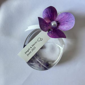 May include: A clear, heart-shaped glass bottle with a purple flower and a pearl bead. A white tag reads "Angel & David June 2025". The bottle is filled with a clear liquid and small purple objects, tied with a white ribbon.
