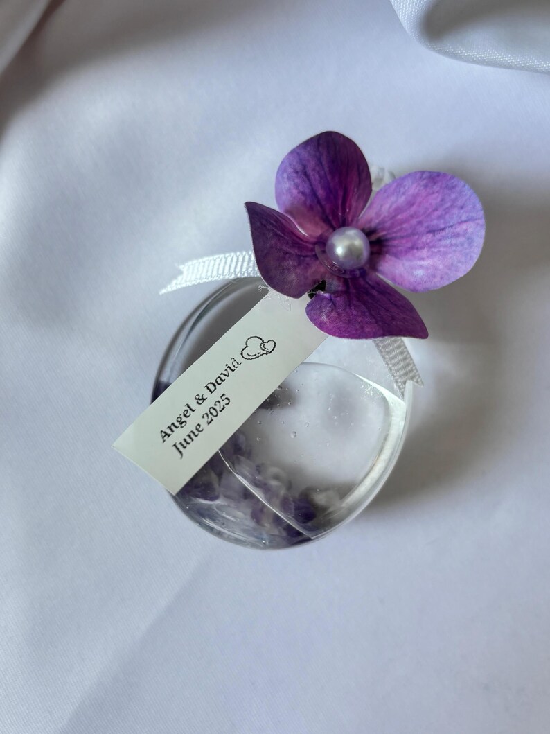 May include: A clear, heart-shaped glass bottle with a purple flower and a pearl bead. A white tag reads "Angel & David June 2025". The bottle is filled with a clear liquid and small purple objects, tied with a white ribbon.