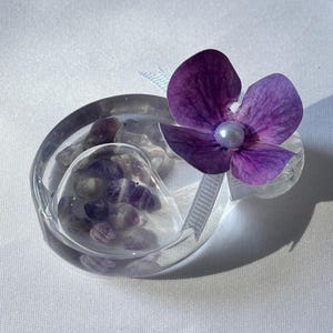 May include: A clear, oval-shaped decorative piece with a heart-shaped compartment filled with small, purple stones. A vibrant purple flower with a pearl center and a white ribbon accent the piece. The item is set against a white background.