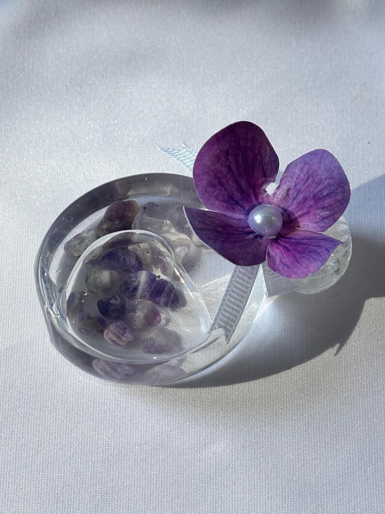 May include: A clear, oval-shaped decorative piece with a heart-shaped compartment filled with small, purple stones. A vibrant purple flower with a pearl center and a white ribbon accent the piece. The item is set against a white background.