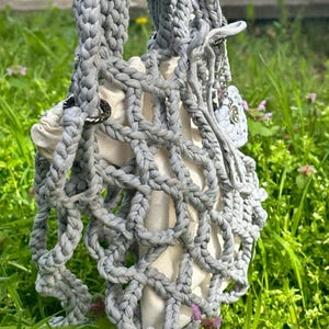 May include: A grey crocheted tote bag with a white lining. The bag has a unique, open-weave design and is held by a hand.