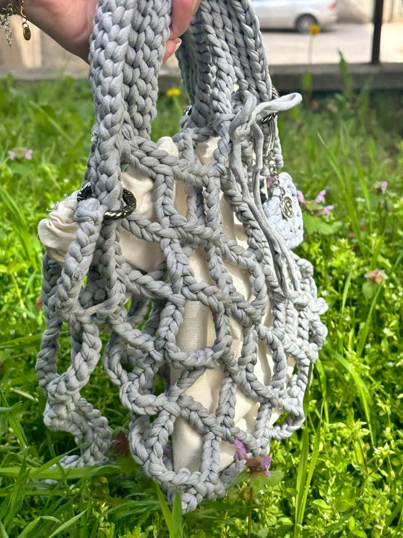 May include: A grey crocheted tote bag with a white lining. The bag has a unique, open-weave design and is held by a hand.